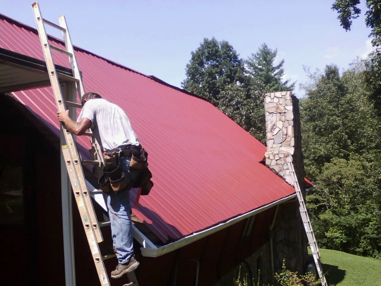 Roofer on a ladder beside a red metal roof and stone chimney — Soffit & Fascia Repair by Roofing Brothers in Shenandoah